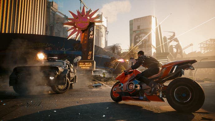 Ein Screenshot aus der Erweiterung Phantom Liberty von Cyberpunk 2077 zeigt, wie der Spieler auf einem roten Motorrad durch die Straßen der Stadt rast, während ein gepanzertes Fahrzeug vor ihm das Feuer eröffnet.